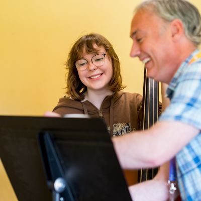 Double bass lesson with tutor in blue checked shirt and student in brown top both smiling and looking at the music smiling